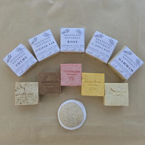 May include: A collection of five Anatolian Naturals handmade soap bars in paper packaging labeled Thyme, Juniper Tar, Rose, St. John's Wort, and Hammam, alongside five unwrapped square soap bars in various earth tones and a round beige loofah sponge.