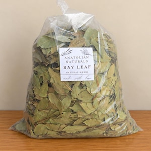 May include: A clear plastic bag filled with dried green bay leaves. A white rectangular label on the front features the text ANATOLIAN NATURALS BAY LEAF NATURAL HERBS made with love, accompanied by small botanical illustrations.