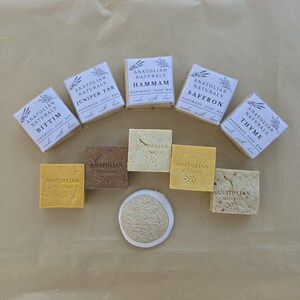 May include: A collection of five square handmade soap bars wrapped in white paper labels with the text Anatolian Naturals, four unwrapped square soap bars in shades of yellow, brown, and beige, and one round textured beige loofah sponge.