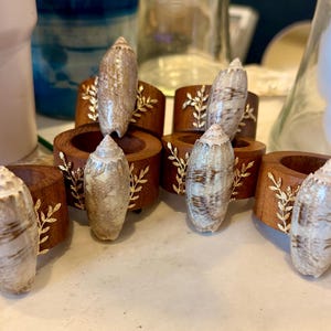 May include: A set of five circular wooden napkin rings, each featuring a polished brown finish. Attached to the front of every ring is a natural, elongated, cream-colored seashell with brown markings, accented by delicate gold-toned metal leaf sprigs.