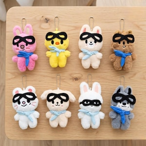 Peut inclure: Collection de huit porte-clés en peluche représentant des animaux de différentes couleurs, dont rose, jaune, blanc et marron. Chaque porte-clés présente un design animalier différent, comme un lapin, un poussin, un chien et un loup, tous portant des masques noirs et des écharpes bleues.