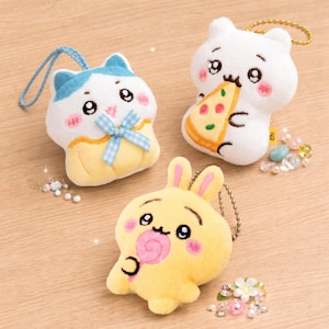 Peut inclure: Trois porte-clés en peluche en forme d'animaux de dessin animé. L'un est un chat blanc avec des détails bleus et un nœud, un autre est un ours blanc mangeant une pizza, et le dernier est un lapin jaune avec une sucette. Chacun a une chaîne en métal.