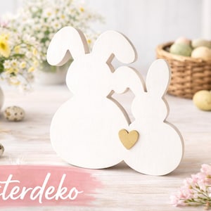 May include: White wooden Easter bunny decoration with a gold heart accent. The decoration is in front of a blurred background with flowers, eggs, and a basket. The word "Osterdeko" is written in pink.