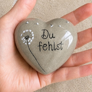Könnte beinhalten: Ein herzförmiger, grauer Stein mit dem deutschen Satz "Du fehlst" in schwarzer Schrift. Auf der linken Seite ist ein weißes Pusteblumen-Design aufgemalt. Der Stein wird in einer Hand gehalten.