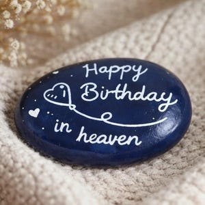 Könnte beinhalten: Ein handbemalter, dunkelblauer Stein mit dem weißen Schriftzug "Happy Birthday in Heaven". Ein herzförmiges Ballon-Design und kleine weiße Herzen akzentuieren den Text. Der Stein liegt auf einem strukturierten, hellfarbigen Stoff.