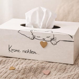 Può includere: Scatola di fazzoletti in legno bianco con illustrazione di gatto nero e cuore dorato. La scatola ha il testo "Krone richten..." e contiene fazzoletti bianchi. Cuori decorativi sparsi.