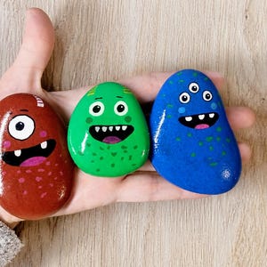 Op de afbeelding: Drie handgeschilderde monsterstenen in rood, groen en blauw. Elke steen heeft een uniek monstergezicht met grote ogen en een lachende mond. De stenen zijn glad en glanzend, perfect als speelgoed of decoratie.