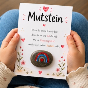 May include: A greeting card with the word "Mutstein" and a message in German, held by a person. The card features a painted stone with a rainbow design. The card is decorated with hearts and flowers.