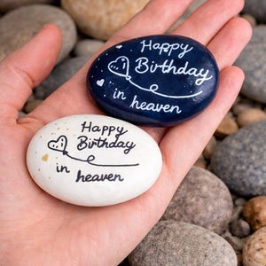 Könnte beinhalten: Zwei bemalte Steine mit der Aufschrift "Happy Birthday in heaven". Ein Stein ist weiß mit goldenen Akzenten, der andere ist dunkelblau. Der Text ist in weißer Schrift mit einem Herz- und Ballon-Design gehalten. Die Steine werden in einer Hand gehalten.
