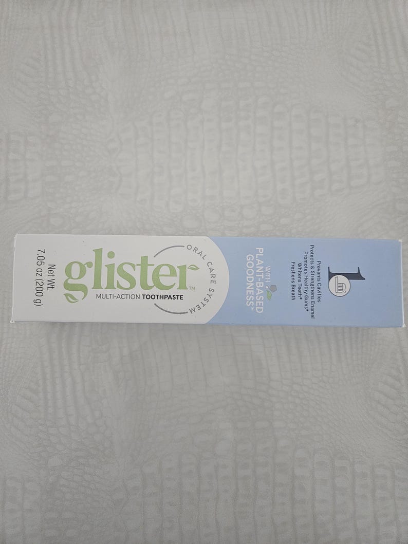 Op de afbeelding: Een tube "glister" multi-action tandpasta. De verpakking is wit met groene en blauwe accenten. De tekst op de tube bevat "Plant-Based Goodness" en "Netto gewicht 200 g". De tandpasta is ontworpen om gaatjes te voorkomen, glazuur te versterken en de adem te verfrissen.