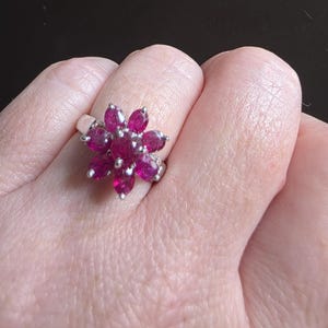 Peut inclure: Une bague en argent ornée d'un motif floral composé de pierres précieuses de couleur rubis, de forme ovale. Les pétales de la bague sont disposés en cercle, créant un motif floral. La bague est portée sur un doigt.