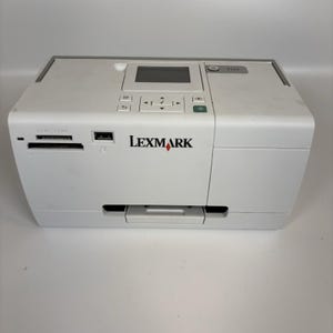 May include: A white Lexmark printer featuring a central digital display screen, a directional navigation keypad, and a power button. The front panel includes a memory card slot and a USB port, with the Lexmark logo printed in black and red.