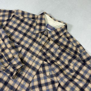 Pode incluir: Uma camisa de manga comprida com botões e um padrão xadrez em tons de azul-marinho, bege e azul-claro. A camisa tem um colarinho pontiagudo, uma carcela de botões frontal e uma etiqueta da marca Pendleton visível no interior do colarinho.
