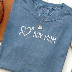 Puede incluir: Una camiseta azul vaquero Comfort Colors con las palabras "BOY MOM" en escritura blanca, con un corazón atravesado por una flecha. Un collar plateado con un colgante de brújula descansa sobre la camisa. Un sombrero marrón está en el fondo.