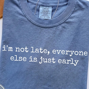 Puede incluir: Camiseta azul vaquero con el texto "i'm not late, everyone else is just early" en blanco. La camiseta tiene cuello redondo y mangas cortas. Se ve un sombrero marrón y un collar plateado.