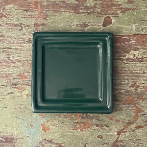 May include: A square, dark green ceramic dish with a glossy finish. The dish has a slightly raised rim and is centered on a distressed, multi-colored wooden surface. The overall aesthetic is rustic and simple.