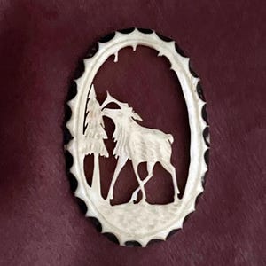 May include: A white carved bone ornament in the shape of an oval with a black border. The ornament depicts a deer standing in front of a tree.