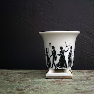 May include: A white ceramic planter with a black silhouette design of people dancing. The planter has a gold trim around the base and a square base.