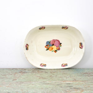 以下が含まれることがあります： 白い楕円形のセラミック製のサービングディッシュで、フルーツのデザインが施されています。デザインは、ピンク、オレンジ、イエロー、ブルーのフルーツが緑の葉で囲まれたものです。