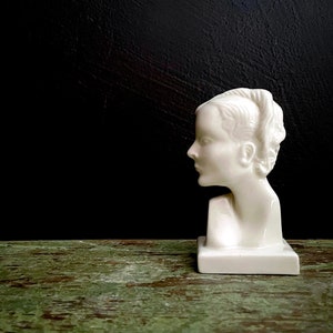May include: White ceramic bust of a woman with her hair styled in a bun. The bust is on a white pedestal and is set against a black background.
