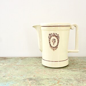 May include: A white ceramic pitcher with a gold-colored design. The design features a stylized letter "P" inside an oval frame with ornate details. The pitcher has a handle on the right side.