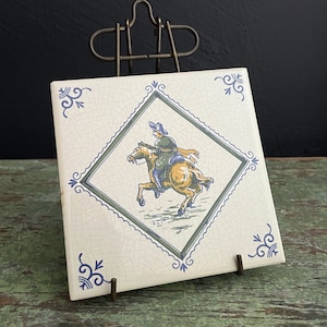 May include: A white ceramic tile with a blue and green border and a diamond-shaped design. The tile features a depiction of a person riding a horse. The tile is displayed on a metal easel.