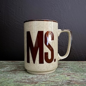 May include: A brown and white speckled ceramic mug with a brown handle. The mug has the letters "MS." printed in brown on the front.