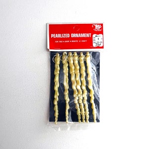 Può includere: Un pacchetto di sette ornamenti a forma di ghiacciolo perlati per un albero, una porta, un camino o un'attività creativa. Gli ornamenti sono di colore giallo pallido e hanno una finitura perlata. La confezione è etichettata "Ornamento perlato" e ha un'etichetta di prezzo di 78 centesimi.