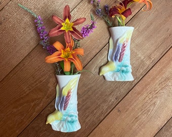May include: Two white ceramic wall vases, each shaped like a boot, are filled with vibrant orange and maroon lilies and purple wildflowers. The vases have colorful painted details, including yellow, blue, and purple accents. The background is a wooden surface.