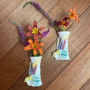 May include: Two white ceramic wall vases, each shaped like a boot, are filled with vibrant orange and maroon lilies and purple wildflowers. The vases have colorful painted details, including yellow, blue, and purple accents. The background is a wooden surface.