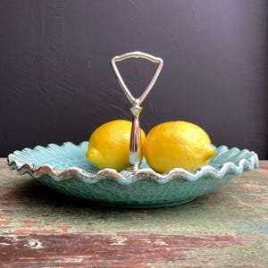 May include: A teal blue ceramic serving tray with a silver handle. The tray has a scalloped edge and is decorated with a textured pattern. Two lemons are sitting on the tray.