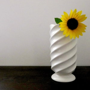 May include: A white ceramic vase with a swirling design, holding a single yellow sunflower with green leaves.
