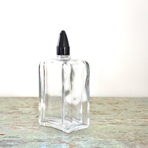 May include: A clear glass bottle with a black plastic stopper. The bottle has a square shape with rounded edges and a textured surface.