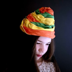 May include: A young person wearing a colorful turban-style hat with orange, yellow, green, and red stripes. The hat is made of fabric and is worn on the head. The person has long brown hair and is looking down.