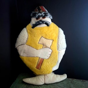 May include: A yellow plush toy shaped like a person wearing a hat and holding an axe. The toy has a white fur trim and a black mustache.