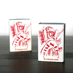 May include: Two red and white matchboxes with a nautical theme. The front of the boxes feature a cartoon illustration of a sailor with a red and white striped shirt and a red hat. The text on the boxes reads "Captain Nemo's Restaurant".
