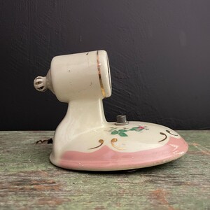 May include: Vintage off-white ceramic lamp with a pink and gold base. The lamp features a floral design and a decorative gold trim. The lamp has a cylindrical head and a curved base. The lamp is an antique and has a unique design.