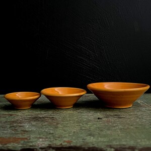 May include: Three orange ceramic bowls of varying sizes, arranged from smallest to largest, on a green wooden surface.