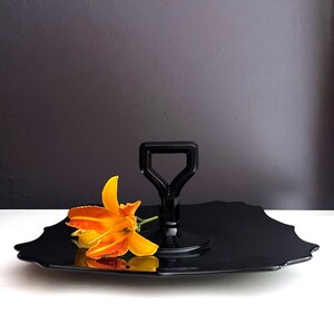 May include: Black glass serving tray with a scalloped edge and a black handle. A single orange flower is resting on the tray.