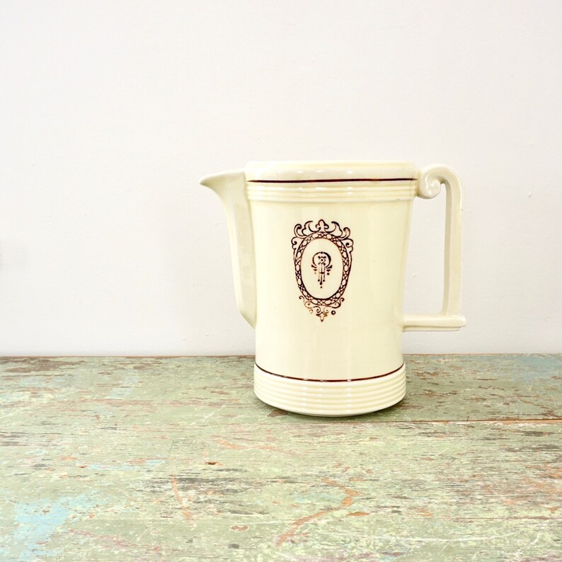 May include: A white ceramic pitcher with a gold-colored design. The design features a stylized letter "P" inside an oval frame with ornate details. The pitcher has a handle on the right side.