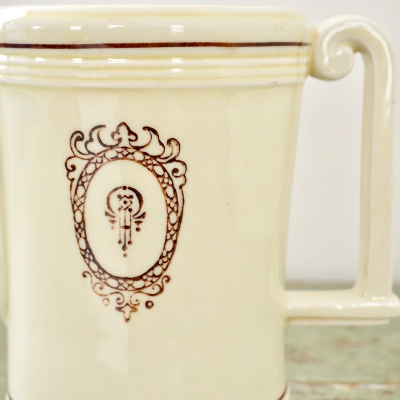 May include: A cream-colored ceramic pitcher with a brown ornate design and two handles. The design features a circle with a stylized symbol inside.