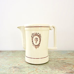 May include: A white ceramic pitcher with a gold-colored design. The pitcher has a wide mouth and a handle on the side. The design features a stylized crest with a keyhole and a decorative frame.