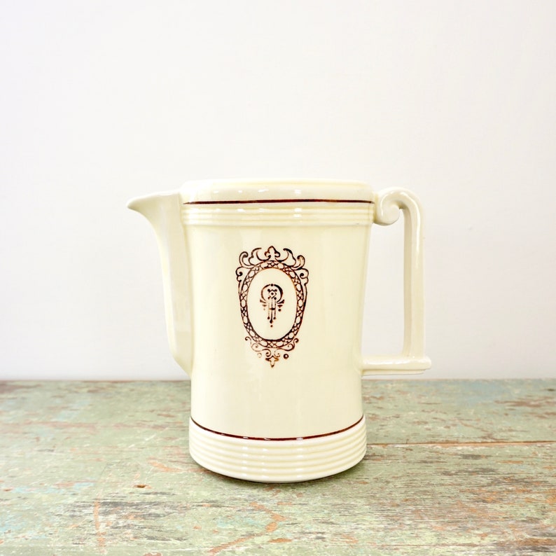 May include: A white ceramic pitcher with a gold-colored design. The pitcher has a wide mouth and a handle on the side. The design features a stylized crest with a keyhole and a decorative frame.