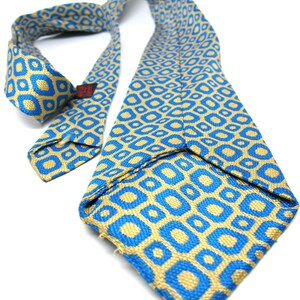 Puede incluir: Una corbata estampada en azul y dorado. La corbata presenta un dise&ntilde;o geom&eacute;trico repetitivo de cuadrados dorados dentro de cuadrados azules. La corbata de tela tejida se muestra sobre un fondo blanco.