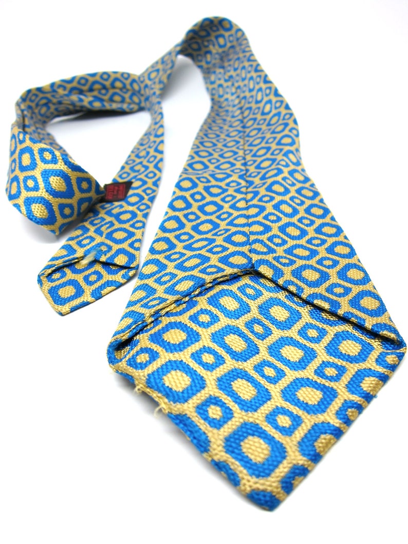 Puede incluir: Una corbata estampada en azul y dorado. La corbata presenta un dise&ntilde;o geom&eacute;trico repetitivo de cuadrados dorados dentro de cuadrados azules. La corbata de tela tejida se muestra sobre un fondo blanco.