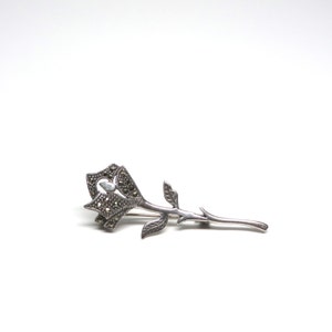May include: A silver rose brooch with a marcasite stone in the center of the flower. The stem is long and slender with leaves on either side.