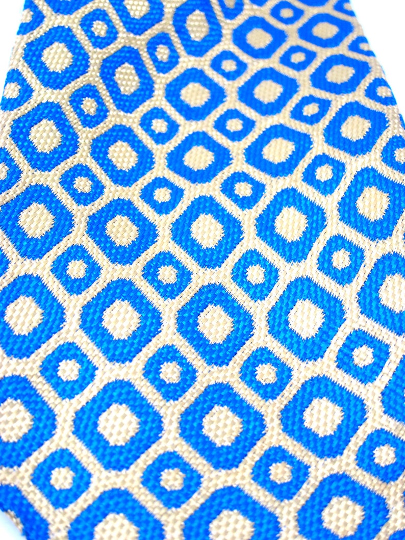 Puede incluir: Primer plano de una corbata con estampado azul y beige. La corbata presenta un dise&ntilde;o geom&eacute;trico repetitivo de formas de diamante azules con centros beige. La tela tiene una apariencia texturizada, lo que sugiere un material tejido. La est&eacute;tica general es moderna y elegante.