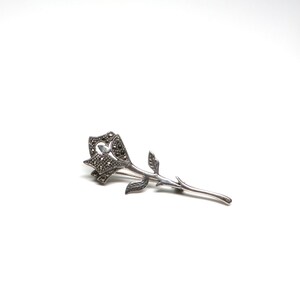 May include: A silver rose brooch with marcasite stones. The rose is detailed with a heart-shaped center and a long stem.