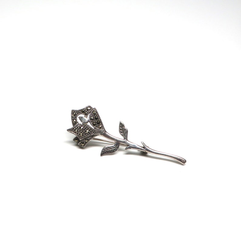 May include: A silver rose brooch with marcasite stones. The rose is detailed with a heart-shaped center and a long stem.