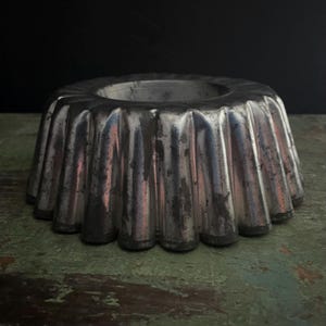 May include: A vintage, fluted aluminum bundt pan. The pan has a circular shape with a central hole and a series of vertical ridges. The metal shows signs of wear, with areas of discoloration and scratches, suggesting age and use. The pan sits on a distressed, green-painted surface.
